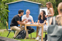 Foto: Vier Personen sitzen an einem Tisch im Freien und diskutieren. Eine Person steht daneben. Im Hintergrund ist ein blaues Gebäude sichtbar. Die Umgebung ist grün und sommerlich.
