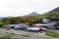 Foto: Eine Biogasanlage in einer ländlichen Umgebung mit grünen Hügeln und herbstlichen Bäumen im Hintergrund. Die Anlage besteht aus mehreren runden Strukturen und einem großen Trichter.
