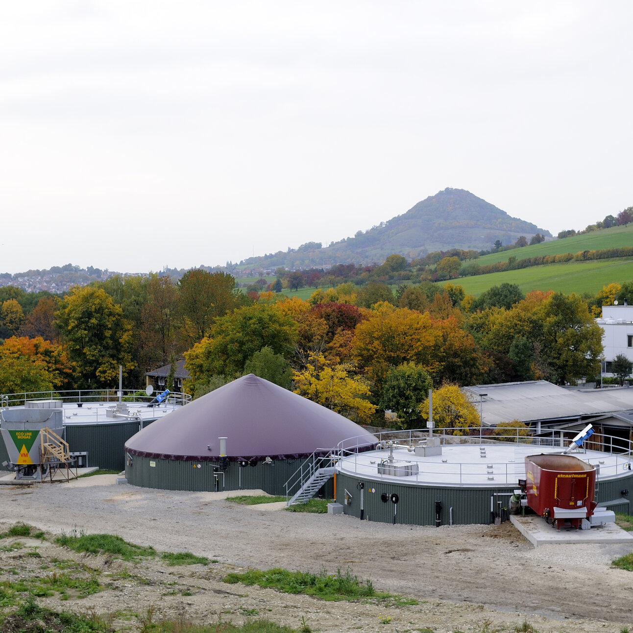 Foto: Eine Biogasanlage in einer ländlichen Umgebung mit grünen Hügeln und herbstlichen Bäumen im Hintergrund. Die Anlage besteht aus mehreren runden Strukturen und einem großen Trichter.