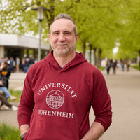 Foto: Eine Person trägt einen roten Hoodie mit der Aufschrift 'Universität Hohenheim' und steht auf einem von Bäumen gesäumten Weg auf dem Campus der Universität Hohenheim. Im Hintergrund sind weitere Personen und Gebäude sichtbar.