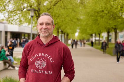 Foto: Eine Person trägt einen roten Hoodie mit der Aufschrift 'Universität Hohenheim' und steht auf einem von Bäumen gesäumten Weg auf dem Campus der Universität Hohenheim. Im Hintergrund sind weitere Personen und Gebäude sichtbar.