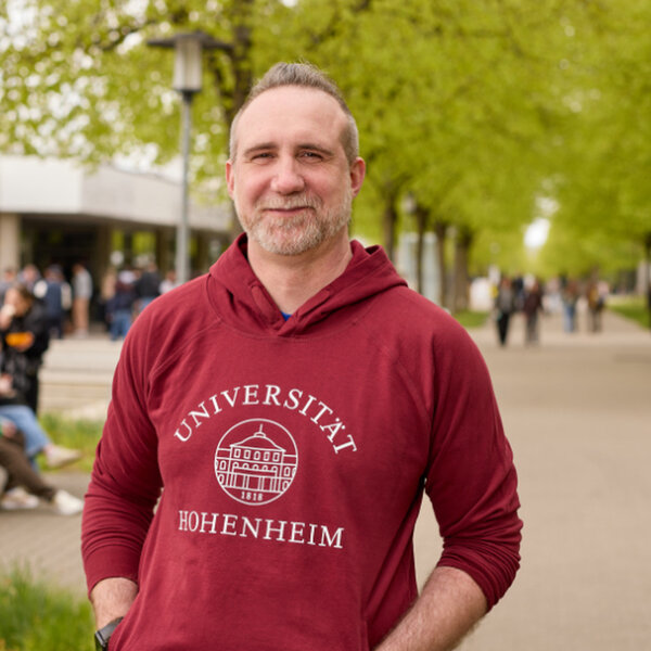 Foto: Eine Person trägt einen roten Hoodie mit der Aufschrift 'Universität Hohenheim' und steht auf einem von Bäumen gesäumten Weg auf dem Campus der Universität Hohenheim. Im Hintergrund sind weitere Personen und Gebäude sichtbar.