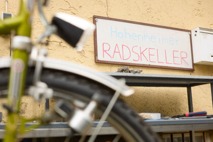 Foto: Ein unscharfes Fahrrad im Vordergrund, dahinter ein Schild mit der Aufschrift 'Hohenheimer RADSKELLER' an einer gelben Wand. Im Hintergrund sind Werkzeuge und Regale sichtbar.