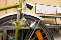 Foto: Ein unscharfes Fahrrad im Vordergrund, dahinter ein Schild mit der Aufschrift 'Hohenheimer RADSKELLER' an einer gelben Wand. Im Hintergrund sind Werkzeuge und Regale sichtbar.