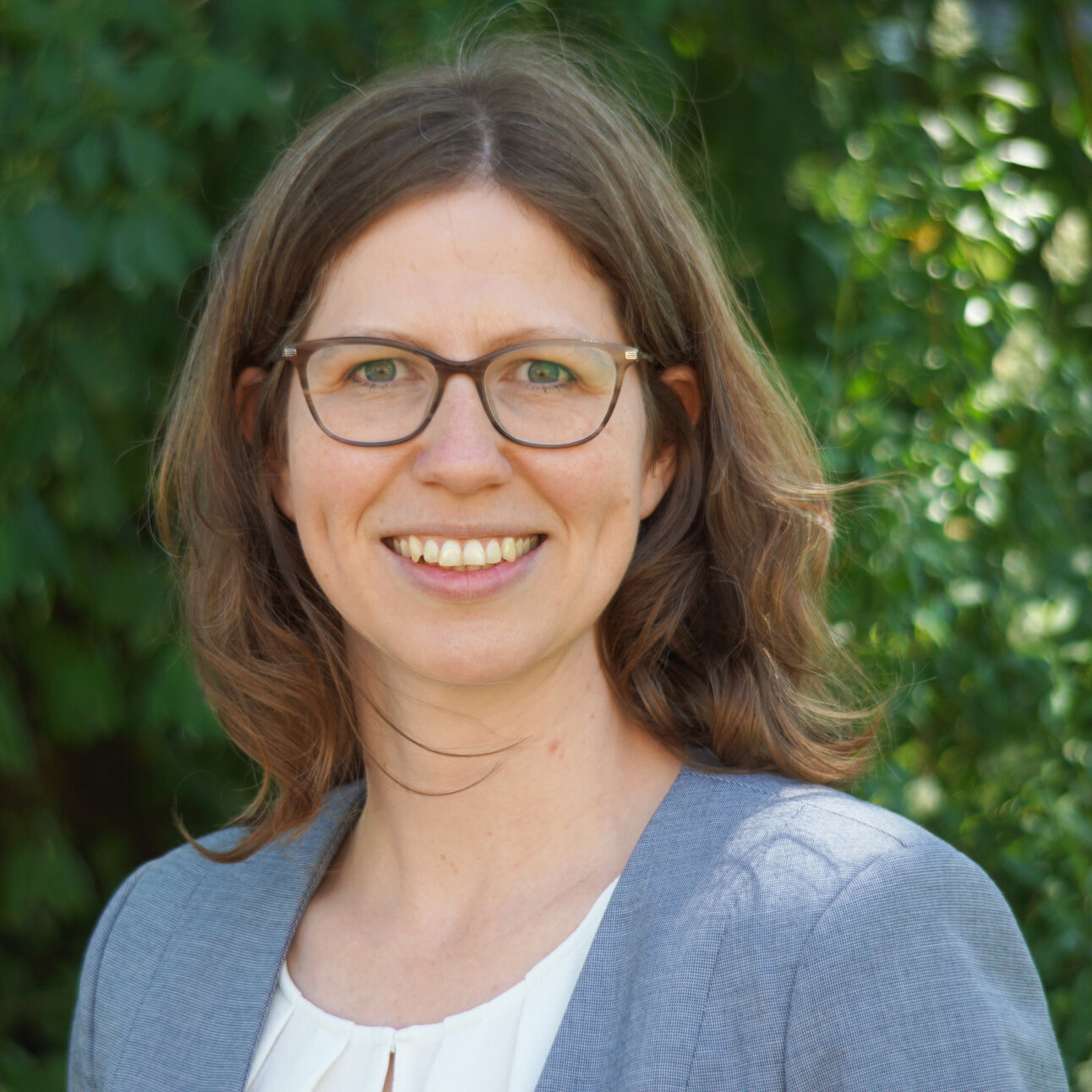 Foto: Eine Person trägt einen grauen Blazer und ein weißes Oberteil, steht im Freien vor einem Hintergrund aus grünen Blättern.