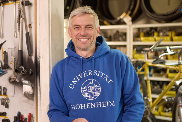 Foto: Eine Person in einem blauen Hoodie mit der Aufschrift 'Universität Hohenheim 1818' steht in einer Werkstatt. An der Wand hängen verschiedene Werkzeuge. Im Hintergrund sind Fahrradteile und Regale sichtbar.