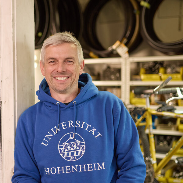 Foto: Eine Person in einem blauen Hoodie mit der Aufschrift 'Universität Hohenheim 1818' steht in einer Werkstatt. An der Wand hängen verschiedene Werkzeuge. Im Hintergrund sind Fahrradteile und Regale sichtbar.