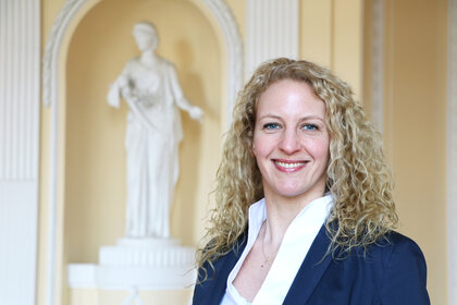 Foto: Eine Person in einem dunklen Blazer steht vor einer weißen Statue in einem Innenraum mit klassischer Architektur.