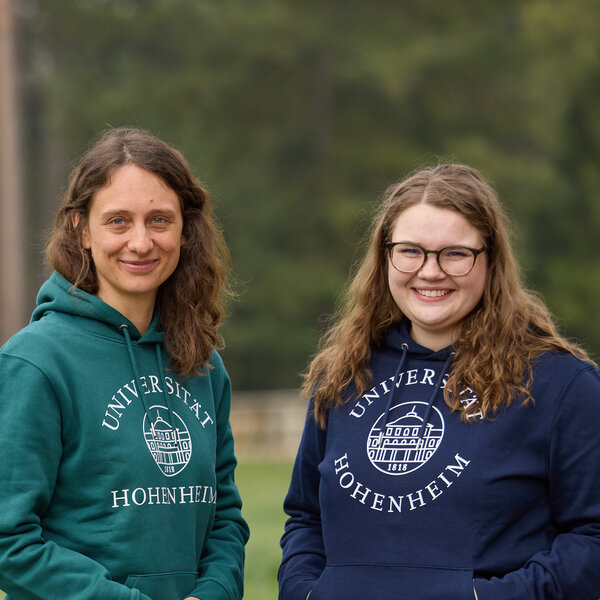 Foto: Zwei Personen stehen im Freien auf einem Rasen. Beide tragen Kapuzenpullover mit dem Logo der Universität Hohenheim. Im Hintergrund sind Bäume und ein Zaun sichtbar.