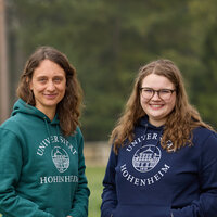Foto: Zwei Personen stehen im Freien auf einem Rasen. Beide tragen Kapuzenpullover mit dem Logo der Universität Hohenheim. Im Hintergrund sind Bäume und ein Zaun sichtbar.