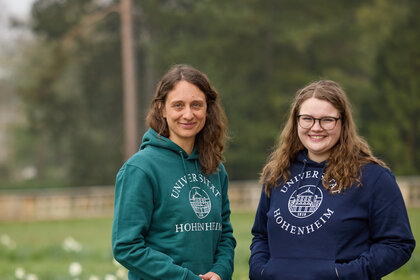 Foto: Zwei Personen stehen im Freien auf einem Rasen. Beide tragen Kapuzenpullover mit dem Logo der Universität Hohenheim. Im Hintergrund sind Bäume und ein Zaun sichtbar.