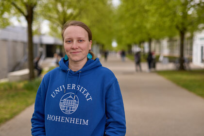 Foto: Eine Person trägt einen blauen Hoodie mit der Aufschrift 'Universität Hohenheim 1818' und steht auf einem Campusweg mit Bäumen im Hintergrund.