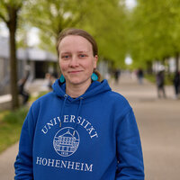 Foto: Eine Person trägt einen blauen Hoodie mit der Aufschrift 'Universität Hohenheim 1818' und steht auf einem Campusweg mit Bäumen im Hintergrund.