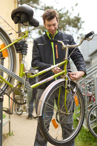 Foto: Eine Person steht draußen und repariert ein grünes Fahrrad. Sie hält ein Werkzeug in der Hand und arbeitet am Vorderrad. Im Hintergrund sind weitere Fahrräder und ein Gebäude sichtbar.