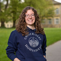 Foto: Eine Person trägt einen blauen Hoodie mit dem Logo der Universität Hohenheim. Im Hintergrund sind zwei weitere Personen auf einem Weg und ein historisches Gebäude sichtbar.