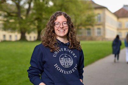 Foto: Eine Person trägt einen blauen Hoodie mit dem Logo der Universität Hohenheim. Im Hintergrund sind zwei weitere Personen auf einem Weg und ein historisches Gebäude sichtbar.