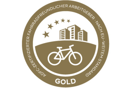 Infografik: Ein goldenes Abzeichen mit einem Fahrrad und Gebäuden, umgeben von Sternen. Text: 'ADFC-Zertifizierter fahrradfreundlicher Arbeitgeber nach EU-weitem Standard'.