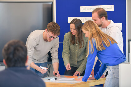 Foto: Eine Gruppe von Personen steht um einen Tisch und arbeitet an einem Projekt. Im Hintergrund sind Schilder mit Texten wie 'Digitalisierung' sichtbar.