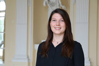 Foto: Eine Person in schwarzer Kleidung steht in einem Innenraum mit klassischen Säulen und einer Skulptur im Hintergrund. Die Umgebung wirkt elegant und formell.
