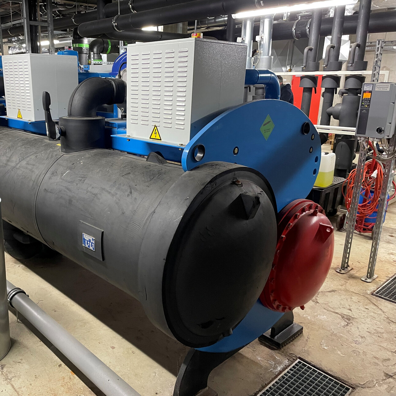 Foto: Eine große industrielle Maschine zur Kälteerzeugung mit mehreren Rohren und Ventilen in einem technischen Raum. Die Maschine ist schwarz mit blauen und roten Elementen. Im Hintergrund sind weitere technische Installationen sichtbar.