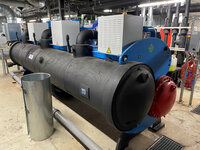 Foto: Eine große industrielle Maschine zur Kälteerzeugung mit mehreren Rohren und Ventilen in einem technischen Raum. Die Maschine ist schwarz mit blauen und roten Elementen. Im Hintergrund sind weitere technische Installationen sichtbar.