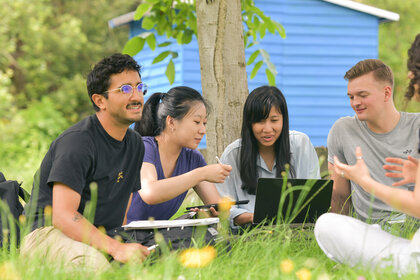 Foto: Eine Gruppe von Personen sitzt im Gras unter einem Baum und arbeitet gemeinsam an einem Laptop. Im Hintergrund ist ein blaues Gebäude sichtbar. Die Umgebung ist grün und natürlich.