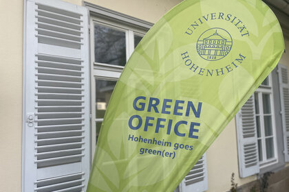 Foto: Ein grünes Banner mit dem Logo der Universität Hohenheim und der Aufschrift 'GREEN OFFICE Hohenheim goes green(er)' steht vor einem Gebäude mit weißen Fensterläden.