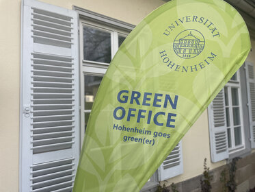 Foto: Ein grünes Banner mit dem Logo der Universität Hohenheim und der Aufschrift 'GREEN OFFICE Hohenheim goes green(er)' steht vor einem Gebäude mit weißen Fensterläden.