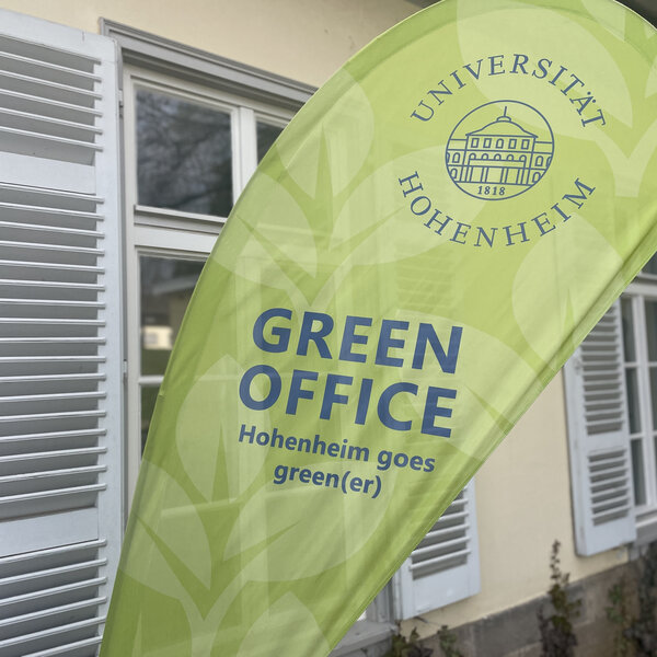 Foto: Ein grünes Banner mit dem Logo der Universität Hohenheim und der Aufschrift 'GREEN OFFICE Hohenheim goes green(er)' steht vor einem Gebäude mit weißen Fensterläden.