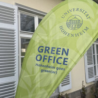 Foto: Ein grünes Banner mit dem Logo der Universität Hohenheim und der Aufschrift 'GREEN OFFICE Hohenheim goes green(er)' steht vor einem Gebäude mit weißen Fensterläden.