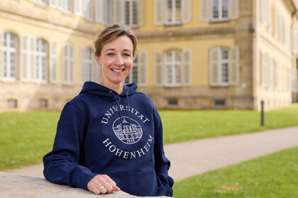 Foto: Eine Person steht vor dem Schloss Hohenheim und trägt einen blauen Hoodie mit dem Aufdruck 'Universität Hohenheim 1818'. Im Hintergrund ist das historische Gebäude mit symmetrischen Fenstern sichtbar.