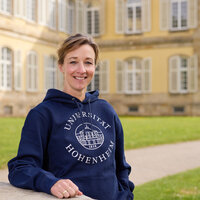 Foto: Eine Person steht vor dem Schloss Hohenheim und trägt einen blauen Hoodie mit dem Aufdruck 'Universität Hohenheim 1818'. Im Hintergrund ist das historische Gebäude mit symmetrischen Fenstern sichtbar.