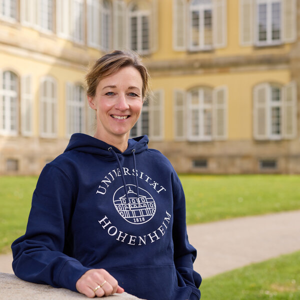 Foto: Eine Person steht vor dem Schloss Hohenheim und trägt einen blauen Hoodie mit dem Aufdruck 'Universität Hohenheim 1818'. Im Hintergrund ist das historische Gebäude mit symmetrischen Fenstern sichtbar.