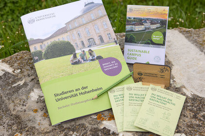 Foto: Verschiedene Broschüren und Flyer der Universität Hohenheim liegen auf einem Stein. Eine Broschüre zeigt das Schloss Hohenheim und trägt den Titel 'Studieren an der Universität Hohenheim'. Ein anderer Flyer ist ein 'Sustainable Campus Guide'.