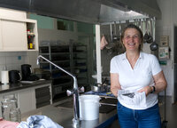 Foto: Eine Person steht in einer Küche neben einer Spüle. Auf der Arbeitsfläche sind Gläser und ein Handtuch zu sehen. Im Hintergrund hängen Küchenutensilien an der Wand.