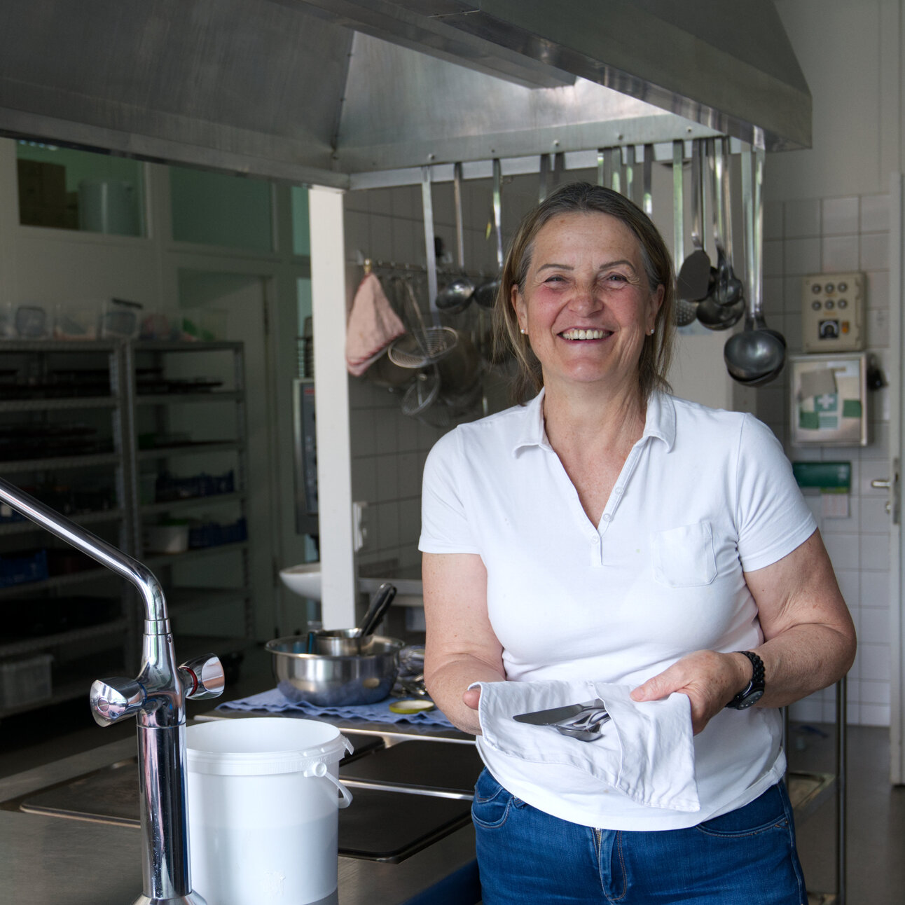 Foto: Eine Person steht in einer Küche neben einer Spüle. Auf der Arbeitsfläche sind Gläser und ein Handtuch zu sehen. Im Hintergrund hängen Küchenutensilien an der Wand.