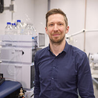 Foto: Eine Person steht in einem Labor mit wissenschaftlicher Ausrüstung im Hintergrund, darunter Flaschen mit blauen Deckeln und technische Geräte.