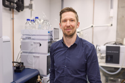 Foto: Eine Person steht in einem Labor mit wissenschaftlicher Ausrüstung im Hintergrund, darunter Flaschen mit blauen Deckeln und technische Geräte.