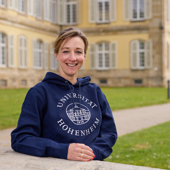 Foto: Eine Person trägt einen blauen Hoodie mit dem Logo der Universität Hohenheim. Im Hintergrund ist das Schloss Hohenheim mit seinen charakteristischen Fenstern und der gepflegten Rasenfläche zu sehen.