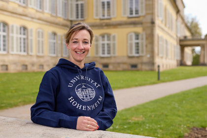 Foto: Eine Person trägt einen blauen Hoodie mit dem Logo der Universität Hohenheim. Im Hintergrund ist das Schloss Hohenheim mit seinen charakteristischen Fenstern und der gepflegten Rasenfläche zu sehen.
