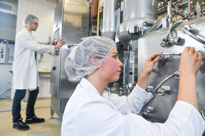 Foto: Zwei Personen in einem Labor, die weiße Kittel und Haarnetze tragen. Eine Person arbeitet an einer großen Edelstahlmaschine, während die andere an einem Bedienfeld steht. Die Umgebung ist industriell mit Rohren und Ventilen.