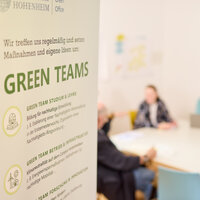 Foto: Ein Poster mit der Aufschrift 'Green Teams' und Text über nachhaltige Bildung und Infrastruktur ist im Vordergrund. Im Hintergrund sitzen Personen an einem Tisch in einem Besprechungsraum der Universität Hohenheim.