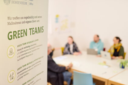 Foto: Ein Poster mit der Aufschrift 'Green Teams' und Text über nachhaltige Bildung und Infrastruktur ist im Vordergrund. Im Hintergrund sitzen Personen an einem Tisch in einem Besprechungsraum der Universität Hohenheim.